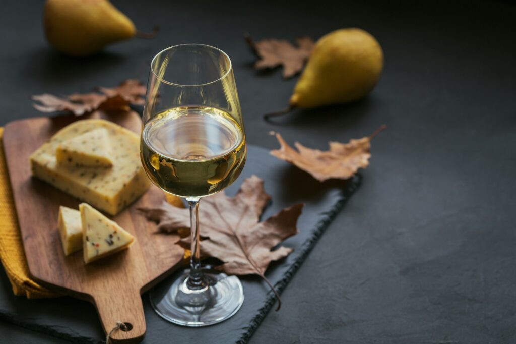 A glass of white wine served with cheese in a cutting board on dark background